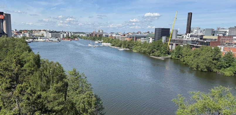 Utsikt över Ulvsundasjön med segelbåtar förtöjda längs stranden, omgiven av lummig grönska. I bakgrunden syns stadsbebyggelse och till höger ett industriområde med en gul lyftkran och ett högt skorstensliknande torn. Himlen är delvis molntäckt med blå partier.